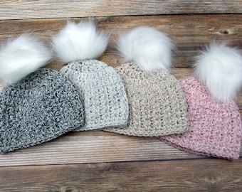 May include: Four crocheted hats with white faux fur pom poms. The hats are in shades of gray, beige, and pink.
