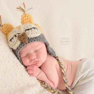 May include: A sleeping baby wearing a handmade owl hat with closed eyes. The hat is primarily gray, cream, and yellow, with braided ear flaps. The baby is lying on a cream-colored blanket. The image includes the text "CARYN Scanlan Newborn".