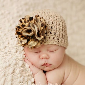May include: A newborn baby wearing a crocheted beige hat with a brown and black leopard print flower on top. The baby is sleeping with their hands tucked under their chin.