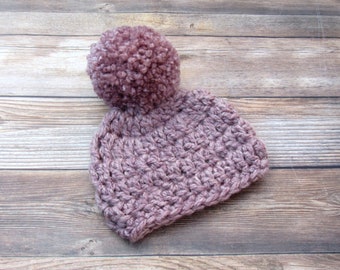 Könnte beinhalten: Eine mauvefarbene Häkelmütze mit einem flauschigen Bommel oben. Die Mütze ist aus weichem Garn gefertigt und hat ein schlichtes, klassisches Design.