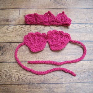 Op de afbeelding: Een fel magenta haakset bestaande uit een hoofdband met een geschulpte rand en een top met twee schelpvormige cups verbonden door een lang, dun gehaakt koord. Het garen heeft een getextureerd, gelust uiterlijk over het hele kledingstuk.