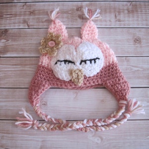 May include: A pink and brown knitted owl hat with a flower on top and a long braided tie. The owl has white eyes and a brown beak.
