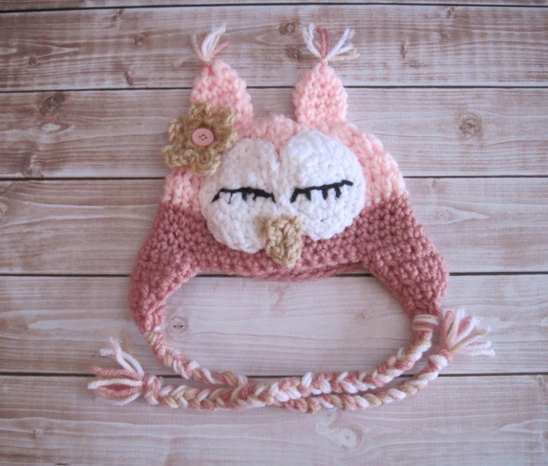 May include: A pink and brown knitted owl hat with a flower on top and a long braided tie. The owl has white eyes and a brown beak.