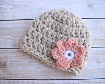 Op de afbeelding: Een beige gebreide baby muts met een roze gebreide bloem erop.