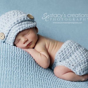 May include: A sleeping baby wearing a light blue crocheted hat with wooden buttons and a matching light blue crocheted diaper. The baby is lying on a light blue textured blanket.