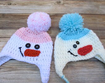 Puede incluir: Dos gorros de muñeco de nieve de crochet hechos con hilo blanco, rosa y azul. Los gorros tienen ojos de botón negro, una nariz roja de crochet y una sonrisa bordada negra. Un gorro tiene un pompón rosa y el otro un pompón azul.