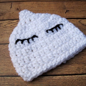 May include: A white crocheted hat with black eyelashes. The hat is handmade and has a soft, textured appearance. The hat is designed to resemble a sleeping face.