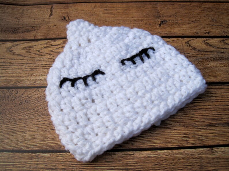 May include: A white crocheted hat with black eyelashes. The hat is handmade and has a soft, textured appearance. The hat is designed to resemble a sleeping face.