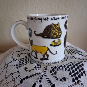 Könnte beinhalten: Ein weißer Keramikbecher mit Illustrationen verschiedener Katzen, einem blauen Ball und einem gelben Futternapf mit der Aufschrift PUSS. Der Text Pussy cat pussy cat where have you been ist in schwarzer Schrift oben und unten aufgedruckt.