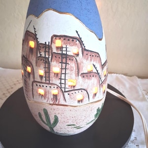 May include: An oval-shaped ceramic lamp with a blue top and a white base featuring a hand-painted desert scene. The design includes brown adobe-style buildings with black ladders and green cacti. Small cutouts emit a warm light from within the lamp.