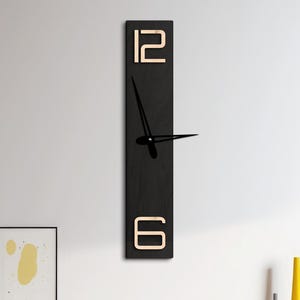 Könnte beinhalten: Eine rechteckige schwarze Wanduhr mit goldfarbenen Ziffern oben und unten, die die Zahlen 12 und 6 anzeigt. Die Uhrzeiger sind schwarz. Der Text "COLOR: WENGE + NATURAL" steht oben.