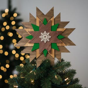 Könnte beinhalten: Ein hölzerner Stern-Baumschmuck mit einem Schneeflocken-Design. Der Stern besteht aus geschichtetem Holz in Braun- und Grüntönen. Die Schneeflocke ist weiß. Der Aufsatz wird auf einen Weihnachtsbaum gesetzt.