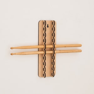 Pode incluir: Um suporte de baquetas de madeira com um design decorativo de borda ondulada. Duas baquetas de madeira são mantidas no lugar pelo suporte. O suporte é montado em uma parede branca.