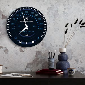 May include: A black and blue Porsche speedometer-style wall clock. The clock face displays a speedometer with the Porsche logo, and the speed is indicated at 227.7 km/h. The clock has white and blue markings and is mounted on a textured wall.