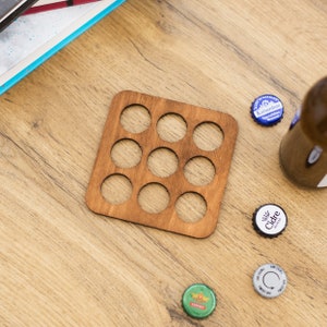 Puede incluir: Un posavasos de madera con nueve recortes circulares para sujetar tapas de botellas. El posavasos es marrón y tiene un acabado de madera natural.