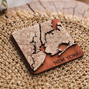 Può includere: Un sottobicchiere in legno con una mappa di New York City incisa al laser. Il sottobicchiere è marrone con una finitura in legno chiaro e il testo "NEW YORK" è inciso nell'angolo in basso a destra.