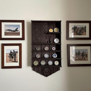 May include: A dark brown wooden display shelf shaped like the American flag, holding military challenge coins. Framed photographs of military personnel and an aircraft are on either side of the shelf.