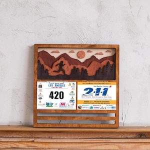 May include: Wooden race bib and medal display featuring a mountain scene and a runner silhouette. Includes race bibs from Los Angeles and San Benito County. The Los Angeles bib has the number 420.