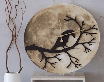 May include: A round wall clock featuring a full moon design with a black bird perched on a tree branch. The clock hands are gold. The clock is displayed on a white shelf with a vase of branches to the left.
