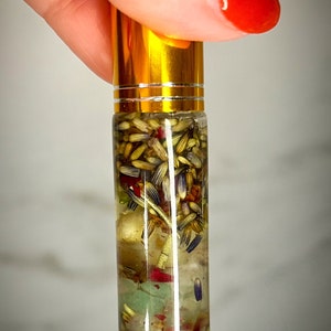 May include: A clear glass roller bottle with a gold lid filled with a yellow liquid and various dried herbs and crystals.