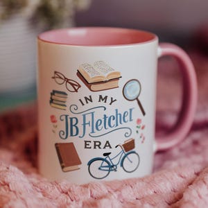 Puede incluir: Taza de cerámica blanca con interior y asa rosa, decorada con ilustraciones temáticas de libros y el texto "IN MY JB Fletcher ERA". El diseño incluye libros, gafas, una lupa y una bicicleta.