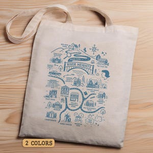 May include: A beige tote bag with a blue illustrated map of River Heights. The map includes landmarks, roads, and text. The bag has two handles and is on a wooden surface. The text "2 COLORS" is at the bottom.