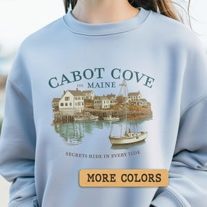 Puede incluir: Sudadera azul claro con una ilustración costera y el texto "Cabot Cove Est. Maine." El diseño muestra una escena portuaria con edificios, barcos y la frase "Secrets Hide in Every Tide." Un botón dice "More Colors."