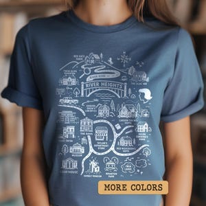 May include: A blue t-shirt with a white illustrated map of River Heights. The map includes landmarks such as the courthouse, department store, and park. The text "MORE COLORS" is in a gold rectangle at the bottom.