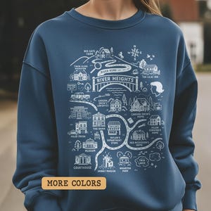 May include: A navy blue sweatshirt featuring a white illustrated map of River Heights. The map includes landmarks like the courthouse, Taylor's Department Store, and Twin Elms Mansion. The words "River Heights" are prominently displayed. A small rectangular button with the words "MORE COLORS" is at the bottom.