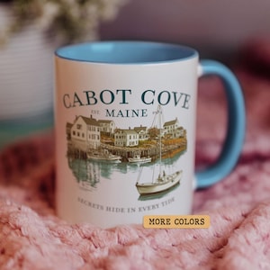 Puede incluir: Taza de cerámica blanca con interior y asa azul claro. La taza presenta una escena costera con las palabras "Cabot Cove Maine" y el texto "Secrets Hide in Every Tide".