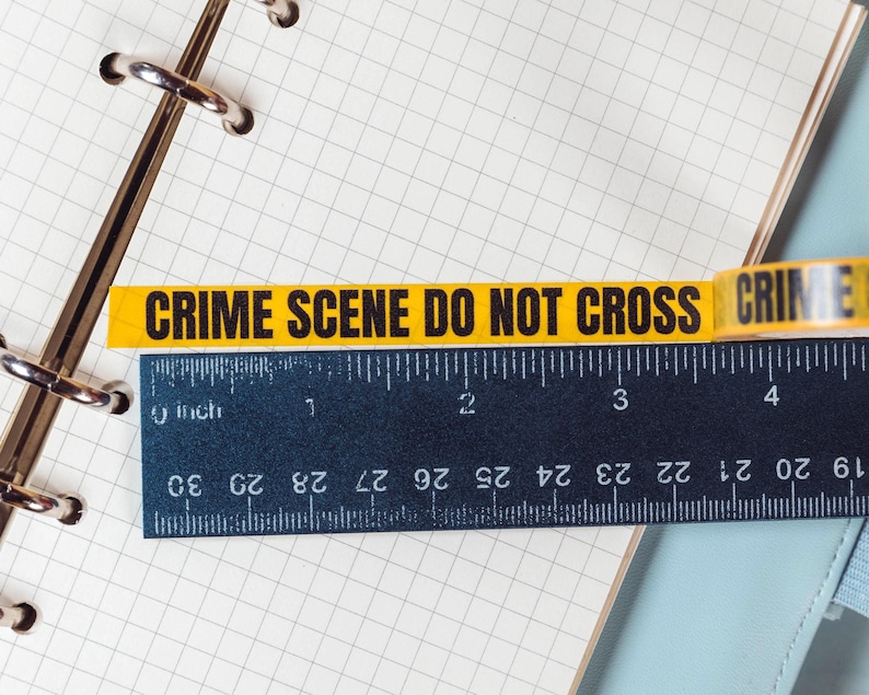 Puede incluir: Una cinta amarilla de escena del crimen con texto negro "CRIME SCENE DO NOT CROSS" se coloca sobre una regla negra con medidas en pulgadas. La regla est&aacute; sobre una p&aacute;gina de cuaderno cuadriculada.