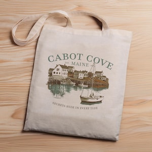 Puede incluir: Una bolsa de lona blanca con un diseño gráfico de un pueblo pequeño con un velero en el puerto. El texto en la bolsa dice "Cabot Cove Est. Maine Secrets Hide in Every Tide".