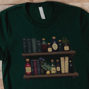 May include: A dark green t-shirt featuring a graphic print of two wooden shelves filled with various vintage books and glass apothecary bottles. The bottles have labels with text including Hemlock, Arsenic, and Morphine, alongside small plants.