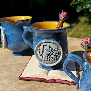 Può includere: Una tazza in ceramica blu con interno giallo, caratterizzata da un medaglione circolare con il testo Tale as old as time. La tazza poggia su un sottobicchiere a forma di libro aperto e presenta una decorazione a rosa scolpita sul manico.