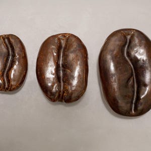 Könnte beinhalten: Drei braune Keramik-Kaffeebohnen in verschiedenen Größen. Die Bohnen sind glatt und glänzend, mit einer realistischen Textur.