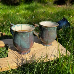 Puede incluir: Dos tazas de cerámica hechas a mano con un esmalte único en tonos turquesa, marrón y crema. Las tazas, llenas de una bebida caliente, descansan sobre una bandeja de madera, rodeadas de hierba verde. Cada taza tiene un asa.