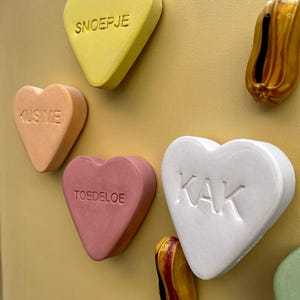 Könnte beinhalten: Eine Sammlung herzförmiger Süßigkeiten mit verschiedenen Botschaften, darunter "KUS ME", "TOEDELOE", "KAK" und "SNOEPJE". Die Süßigkeiten sind aus weißem, rosa und gelbem Material gefertigt und auf einem gelben Hintergrund angeordnet.