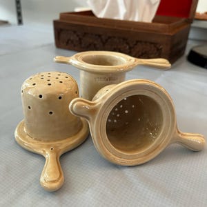 Może przedstawiać: Trzy ceramiczne zaparzacze do herbaty z uchwytami. Zaparzacze są beżowe i mają okrągły kształt z sitkiem na dole. Zaparzacze znajdują się na jasnobrązowej powierzchni.