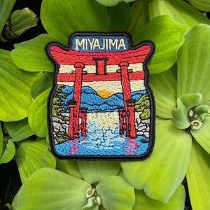 May include: Embroidered patch featuring a red torii gate with the word "MIYAJIMA" above. The design includes a sunset over water and mountains, with a black border. The patch is set against a background of green aquatic plants.