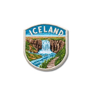 Puede incluir: Un parche bordado en forma de escudo que muestra un paisaje de una cascada que cae sobre acantilados rocosos marrones hacia un r&iacute;o azul. La palabra ICELAND est&aacute; bordada en letras may&uacute;sculas blancas sobre el cielo azul con un borde blanco.