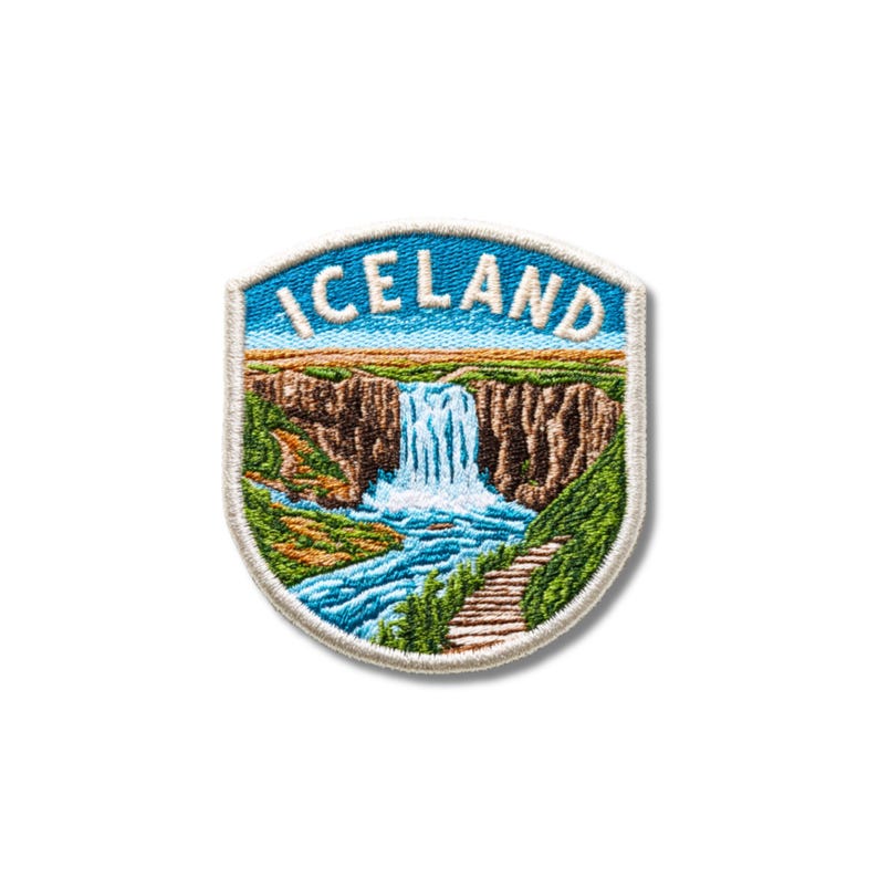 Puede incluir: Un parche bordado en forma de escudo que muestra un paisaje de una cascada que cae sobre acantilados rocosos marrones hacia un r&iacute;o azul. La palabra ICELAND est&aacute; bordada en letras may&uacute;sculas blancas sobre el cielo azul con un borde blanco.