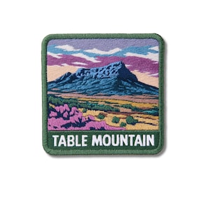 Op de afbeelding: Een vierkante geborduurde patch met een landschap van de Tafelberg met een donkerblauwe top, een goudgele voorgrond en paarse wilde bloemen. De lucht is gevuld met lichtblauwe en paarse wolken. De tekst TABLE MOUNTAIN is in het wit.