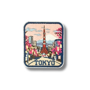 May include: A rectangular embroidered patch featuring a Tokyo cityscape with the red and white Tokyo Tower, pink cherry blossoms, and a mountain in the background. The word TOKYO is stitched in dark blue capital letters at the bottom.
