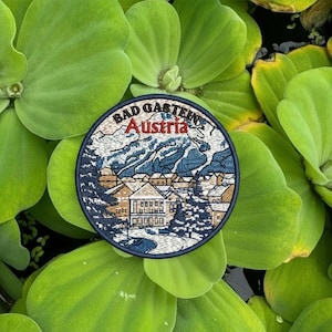 Könnte beinhalten: Ein runder, gestickter Aufnäher mit dem Text "BAD GASTEIN Austria". Der Aufnäher zeigt eine Berglandschaft mit Gebäuden und Bäumen in Blau, Braun und Weiß, vor einem dunkelblauen Rand. Der Aufnäher ist von grünen Blättern umgeben.