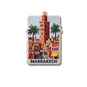 May include: A rectangular embroidered patch featuring a detailed scene of the Koutoubia Mosque tower in Marrakech, surrounded by palm trees and buildings in shades of terracotta, pink, and green, with the word MARRAKECH stitched in blue at the bottom.