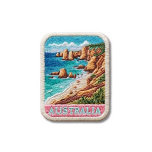 Peut inclure: Un écusson brodé rectangulaire représentant un paysage côtier avec des falaises beiges, une plage de sable et une mer bleue. La scène inclut de la végétation verte et un ciel bleu clair avec des nuages blancs. Le mot AUSTRALIA est brodé en bleu clair.