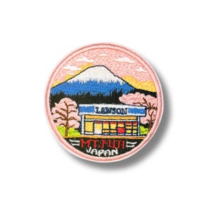 May include: A circular embroidered patch featuring a Lawson convenience store situated in front of a snow-capped Mount Fuji. The design includes pink cherry blossom trees, a blue sky, and the text LAWSON, MT. FUJI, and JAPAN in black lettering.