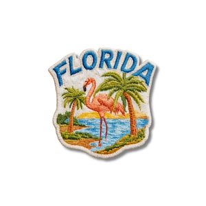 Puede incluir: Un parche bordado que muestra un flamenco rosa de pie en agua azul entre dos palmeras en una orilla arenosa. La palabra FLORIDA está cosida en letras mayúsculas azules en la parte superior del fondo de tela blanca en forma de escudo.