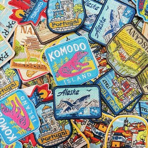 May include: A collection of embroidered travel patches featuring various destinations including Komodo Island with a pink lizard, Alaska with a whale, Portugal with a castle, Lisbon with a yellow tram, and Toronto with a skyline, all in vibrant colors.