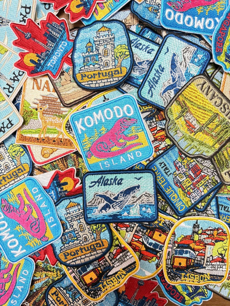May include: A collection of embroidered travel patches featuring various destinations including Komodo Island with a pink lizard, Alaska with a whale, Portugal with a castle, Lisbon with a yellow tram, and Toronto with a skyline, all in vibrant colors.